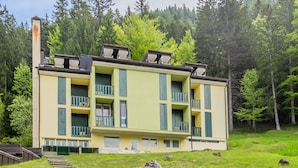 Ein mehrstöckiges Gebäude mit Balkonen, eingebettet in eine bewaldete Landschaft mit einer sanften, grasbewachsenen Böschung davor.