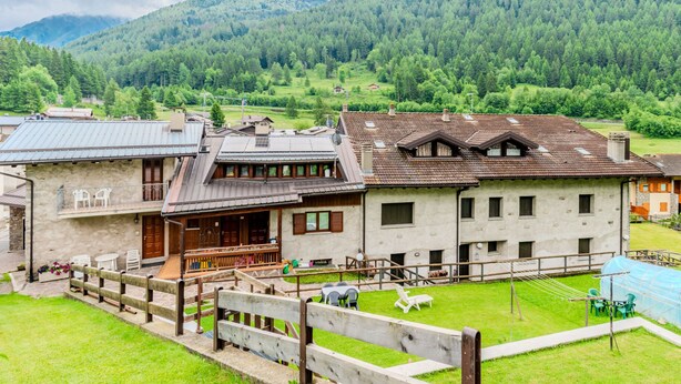 Una construcción rural con terraza de madera, un pequeño jardín y vistas a una cadena montañosa boscosa.