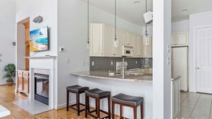 Eine moderne Küche mit weißen Schränken, einer Marmorarbeitsplatte und einem eingebauten Backofen. Drei hölzerne Barhocker und Pendelleuchten befinden sich über der Arbeitsfläche.