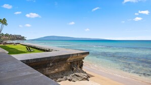 Une plage avec une eau d’un bleu limpide, une structure en béton et une zone herbeuse avec un fauteuil de relaxation.