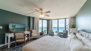 Une chambre d’hôtel avec un grand lit, un bureau avec un ordinateur, deux fauteuils, une petite table avec une lampe, et une vue sur l’océan grâce à de grandes fenêtres.