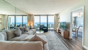 Un salon moderne avec un grand canapé, une table basse et un téléviseur. Il y a un balcon avec vue sur l’océan.