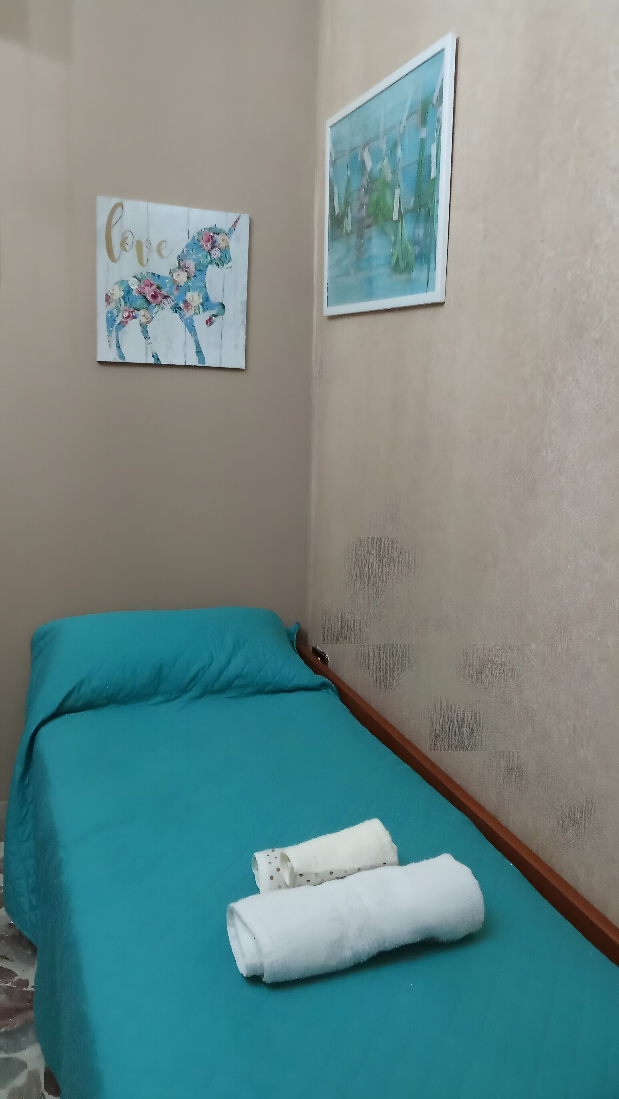 Ein Einzelbett mit türkisfarbener Bettwäsche, ein weißes Handtuch und ein Bild mit floralem Dekor an der Wand.
