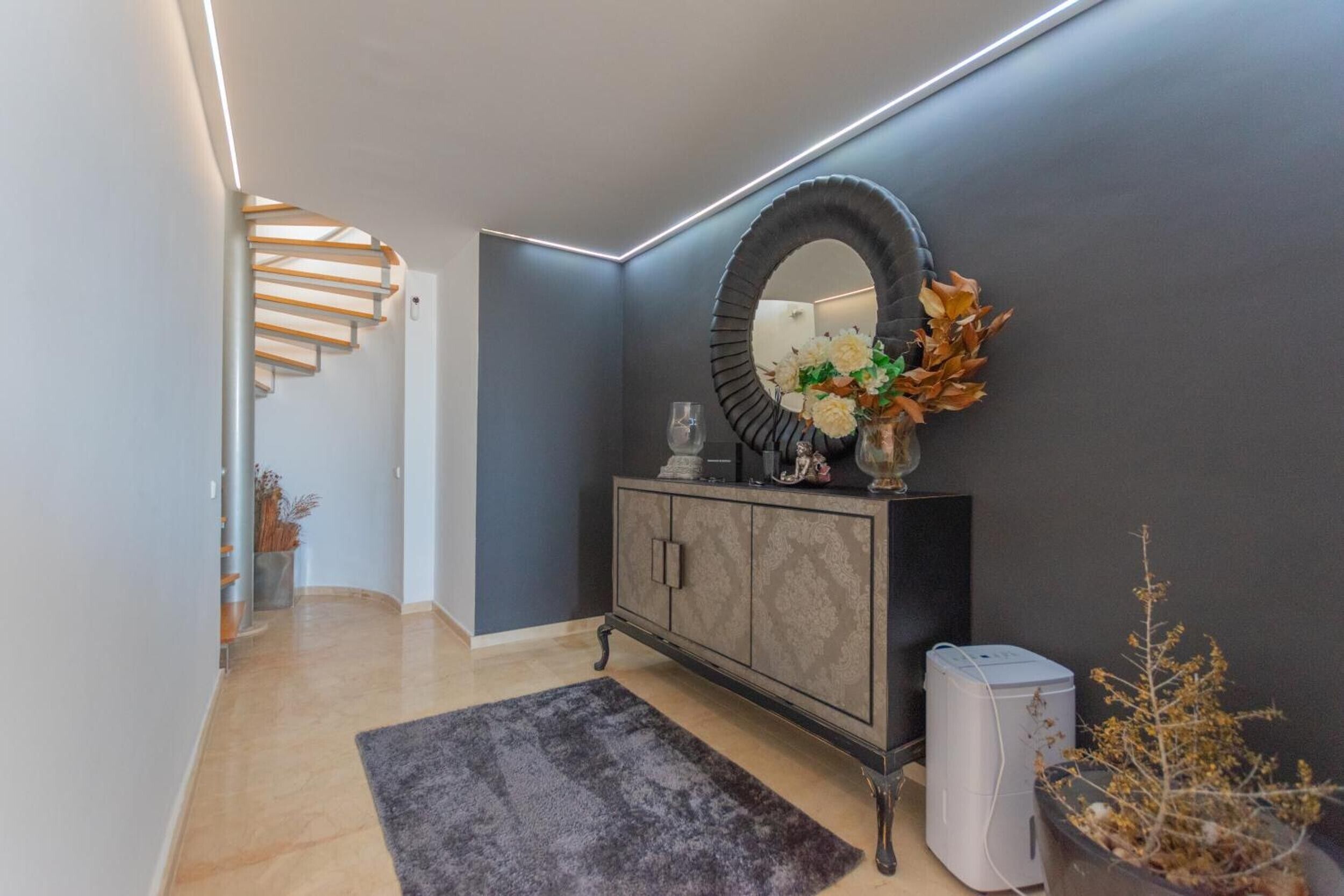 A modern hallway with a dark wall, a round mirror, a cabinet with decorative items, and a staircase.