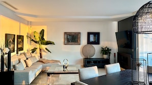 A modern living room with a sofa, coffee table, and dining table. There are potted plants, artwork on the walls, and a TV.