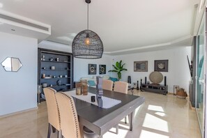 Een eetruimte met een donkere tafel, beige stoelen, een hanglamp en een boekenkast met decoratieve objecten.