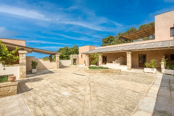 A courtyard with stone flooring, potted plants, and a building with a flat roof and columns.