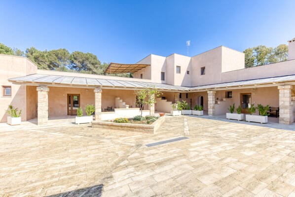 A courtyard with a tiled floor, a covered seating area, and a building with a flat roof and stone columns.