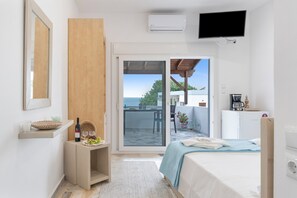A hotel room with a bed, a TV, a minibar, a sliding glass door to a balcony, and a small table with a bottle and glasses.