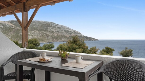 Un balcon avec une table, des chaises, une tasse blanche, une petite plante et une vue sur la mer et les montagnes.
