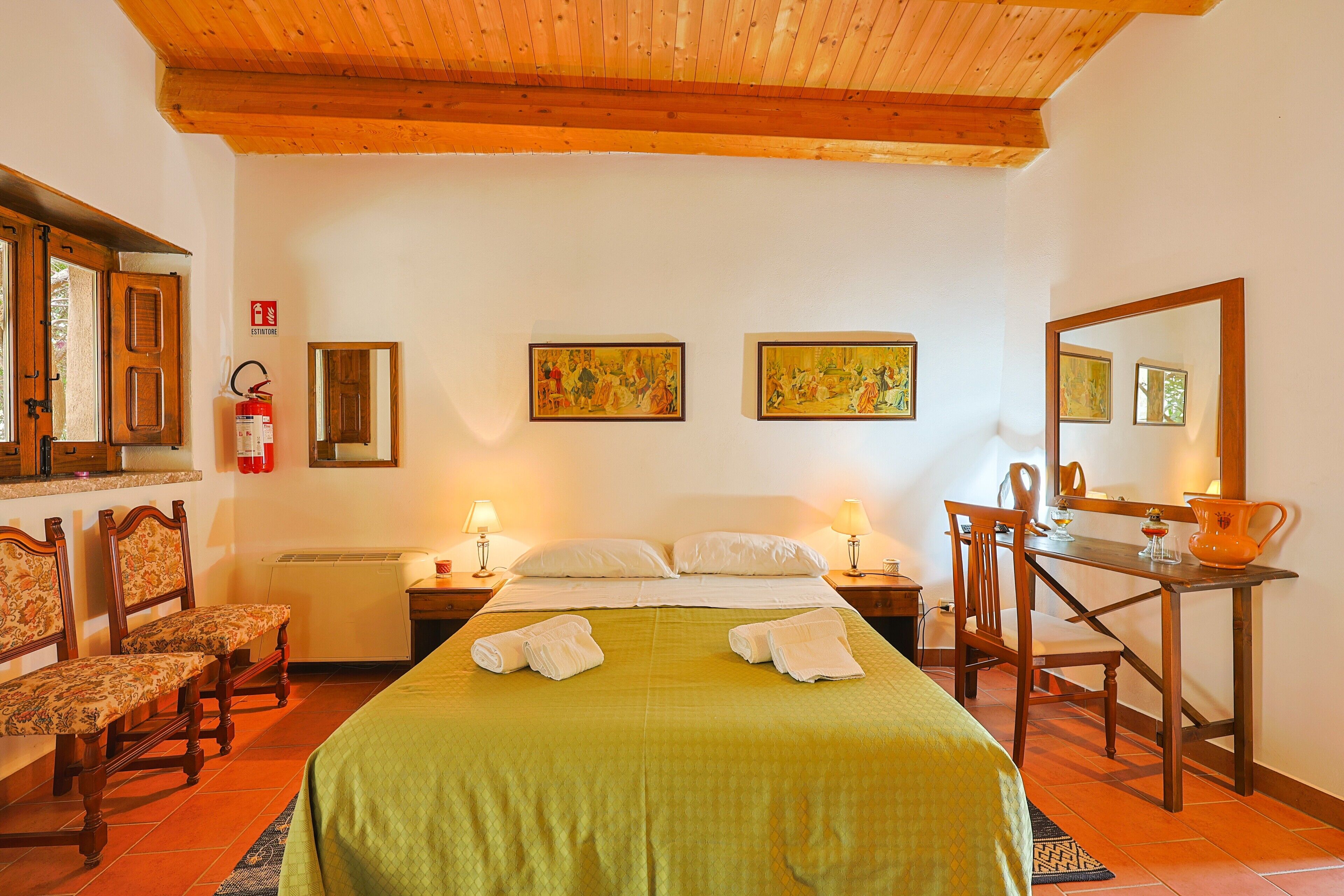 A hotel room with a bed, two chairs, a desk, a mirror, and artwork on the walls.