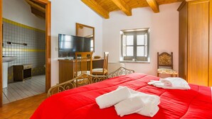 A bedroom with a red bedspread, wooden ceiling beams, a television, a window, and a bathroom with a tiled wall. - 'Writer's Room' with Balcony (San Marco D'alunzio)