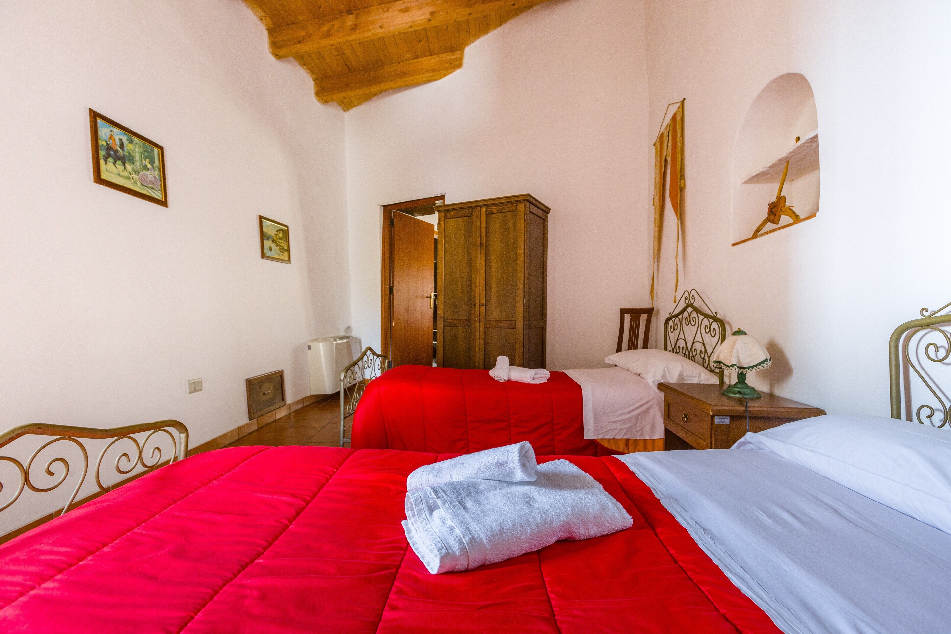 A bedroom with two beds, a wooden wardrobe, a nightstand with a lamp, and a painting on the wall.