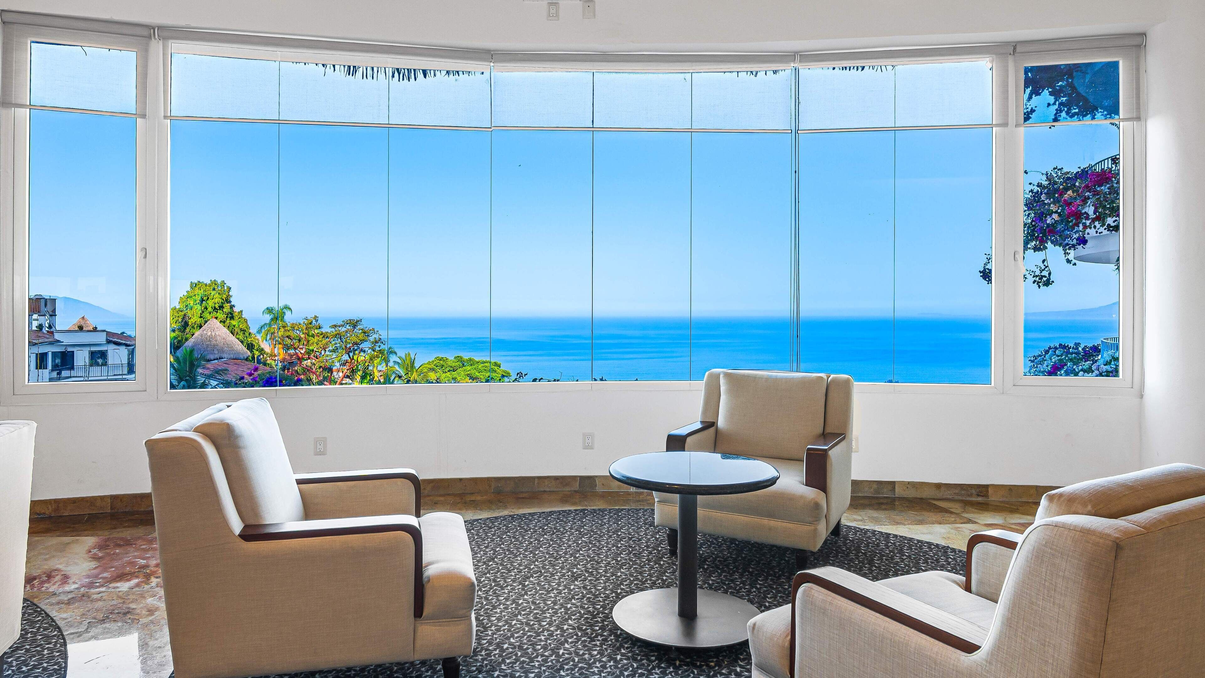 A room with a large panoramic window offering a view of the sea and a tropical landscape.