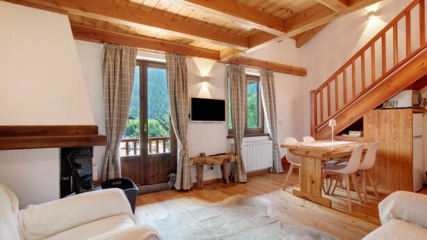 Una habitación acogedora con techo de madera, un televisor montado en la pared, una mesa de comedor con sillas y una escalera.