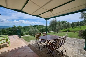 Een overdekt terras met een tafel en stoelen, een groene bank en uitzicht op een groen landschap met bomen en heuvels.