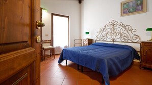 A bedroom with a wooden door, a bed with a blue bedspread, a chair, a bedside table, and a wall-mounted lamp. - Holiday Home 'La Carraia' with Private Terrace, Wi-Fi and Air Conditioning (Suvereto)