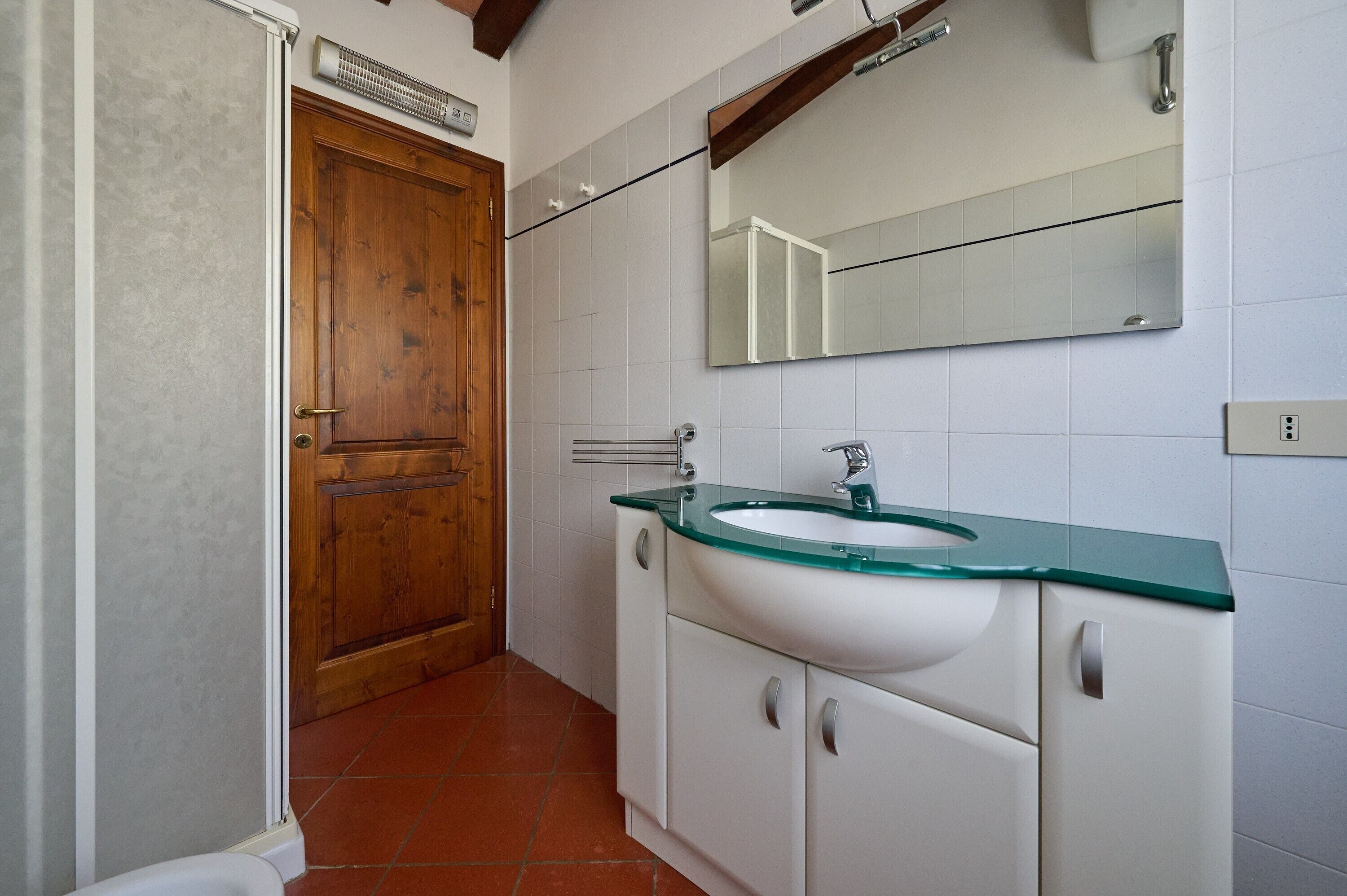 Um banheiro com porta de madeira, paredes com azulejos brancos, piaça branca com bancada verde e um espelho acima da piaça.