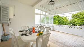 A covered patio with a dining table set, white chairs, and a potted plant.