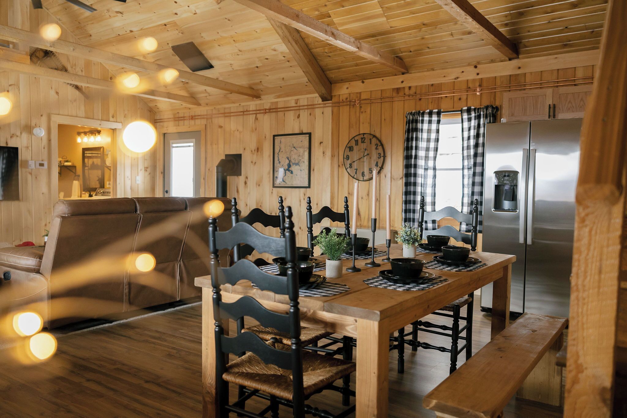 A wooden interior with a dining table, chairs, a clock, and a refrigerator.