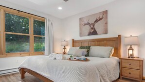 Une chambre avec une grande fenêtre, un lit en bois, deux tables de chevet avec des lampes et un tableau représentant un cerf accroché au mur.