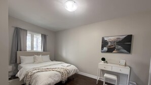 Un dormitorio con cama, mesita de noche, silla, escritorio, un cuadro en la pared y ventana con cortinas.