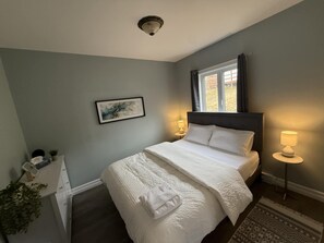 Ein Schlafzimmer mit Bett, Nachttischlampen, einem Fenster, einem gerahmten Bild an der Wand und einem kleinen Schreibtisch mit Pflanze.