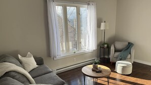 Una sala de estar con un sofá gris, una mesa redonda, una mesita de noche con una lámpara y un sillón con una manta.