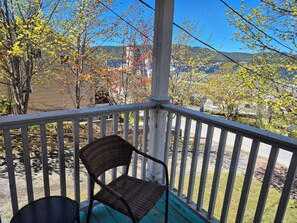 A porch with a wicker chair and a table, overlooking a residential area with trees and a distant view of hills.