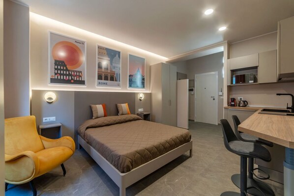 A modern hotel room with a bed, a desk, a chair, and a kitchen area.