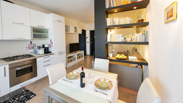 Una cocina moderna con armarios blancos, una mesa de comedor para dos personas y una estantería con libros y objetos decorativos.