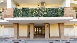 Una entrada de edificio con un camino de acceso cerrado por una reja, un área de acceso cubierta y un balcón con una enredadera verde.