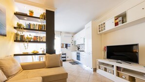 Una sala de estar moderna con un sofá, televisor, estantería y cocina al fondo.