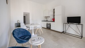 Een moderne keuken met witte kasten, een zwarte tv op een standaard en een eethoek met een blauwe stoel en een kleine ronde tafel.
