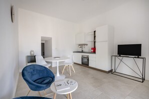 Une cuisine moderne avec des meubles blancs, un téléviseur noir posé sur un meuble, et un coin repas avec une chaise bleue et une petite table ronde.