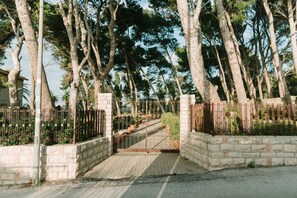Un cancello chiuso da una muratura in pietra, un cancello in metallo e un vialetto lastricato che si inoltra in una zona boschiva.