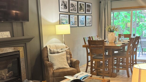 Un salon chaleureux avec une cheminée, des sièges confortables, un coin repas et une grande fenêtre donnant sur un paysage verdoyant.