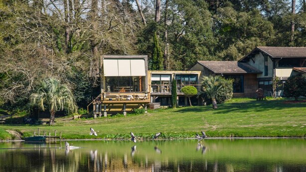 A lakeside property with a wooden deck, a boat, and a reflection of the buildings in the water. - Les Nymphéas, Landes haven of peace, nature & private pond (Angresse)