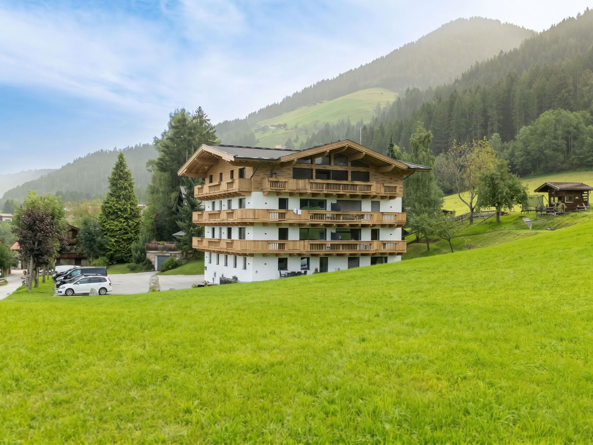 Una chaleta de varios pisos con balcones, ubicada en una zona montañosa con un campo verde al frente.