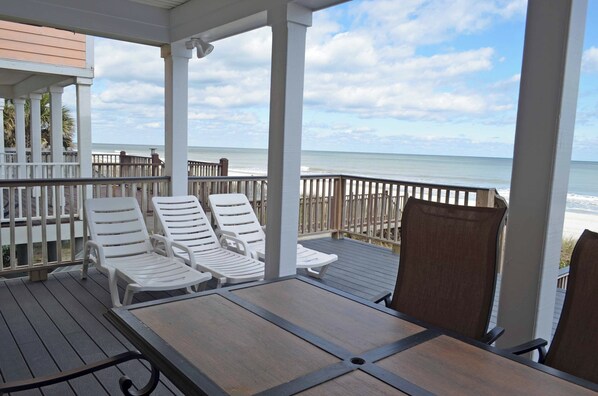A covered deck with white lounge chairs and a table, overlooking the ocean.