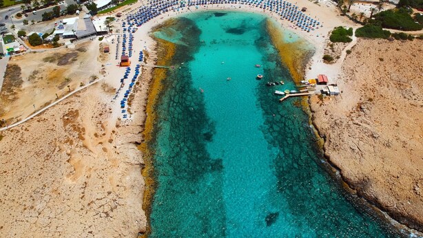 An aerial view of a beach with rows of umbrellas and beach chairs, a pier extending into the water, and buildings in the background.