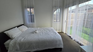A bedroom with a bed, two windows with sheer curtains, a view of a backyard, and a small table. -  Serenity Home 7 by Hiverooms (Pakenham)