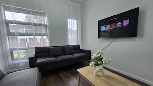 A modern living room with a dark grey sofa, a wooden coffee table with a vase of flowers, and a flat-screen TV mounted on the wall displaying a Netflix interface. -  Serenity Home 7 by Hiverooms (Pakenham)