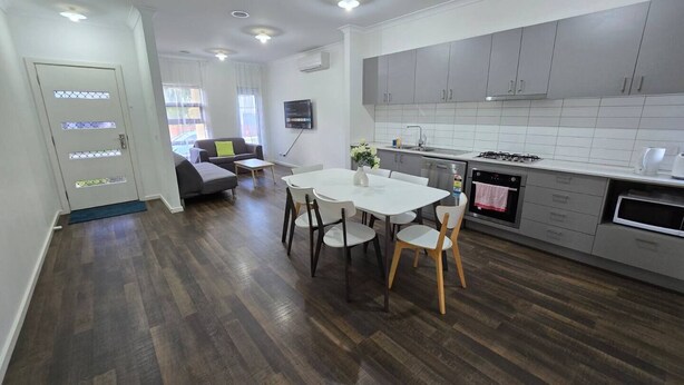 A modern kitchen with grey cabinets, a dining area with a white table and chairs, and a living room with a grey sofa and a TV. -  Serenity Home 7 by Hiverooms (Pakenham)