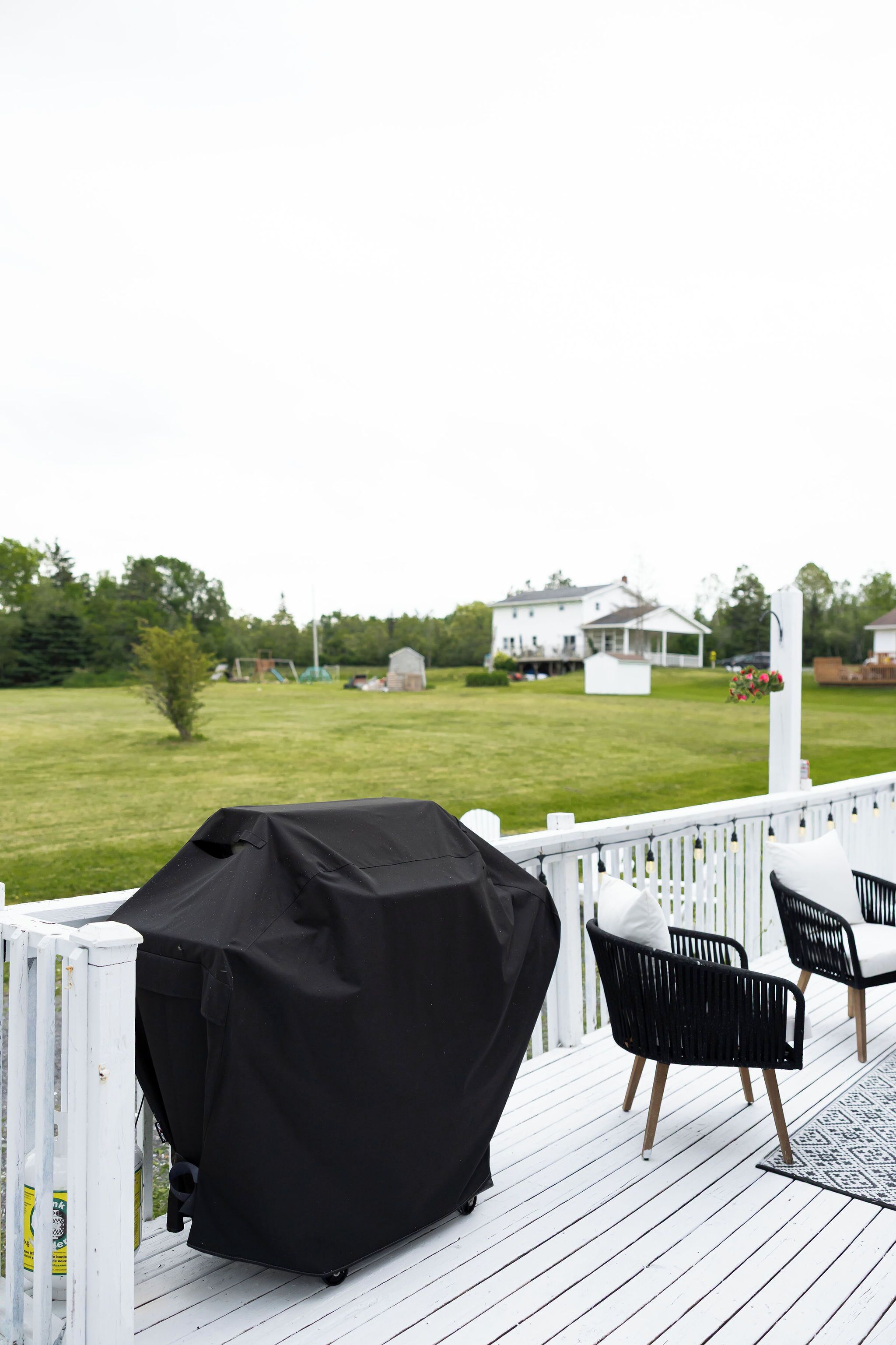 A covered grill on a deck with chairs and a view of a white house in the background.