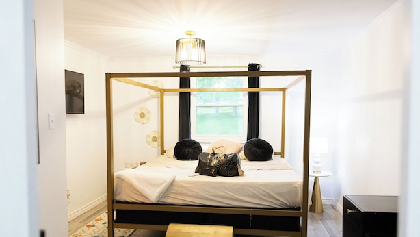 Een hemelbed met gouden frame, een nachtkastje, een lamp en uitzicht op het omliggende landschap door een raam.