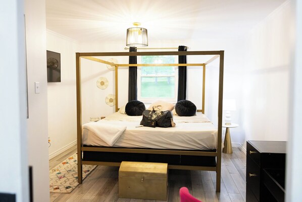 A four-poster bed with a gold frame, a bedside table, a lamp, and a view of the outdoors through a window.