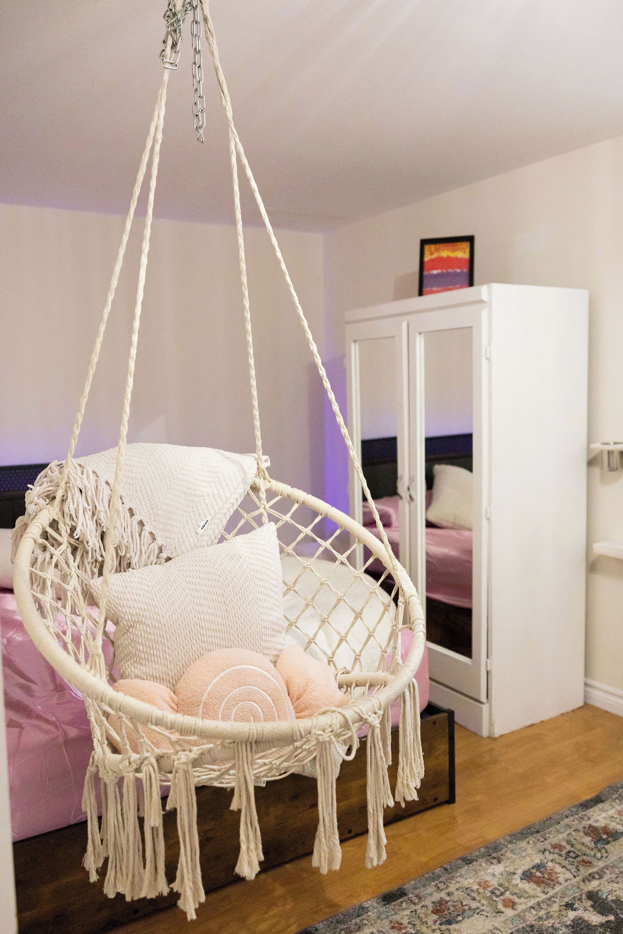 Une pièce avec un fauteuil suspendu en macramé, une armoire et un lit avec une literie rose.