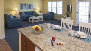 A living room with a blue sofa set, a coffee table, and a kitchen area with a granite countertop, a bowl of fruit, and a knife.