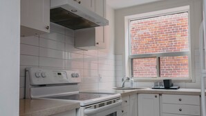 A kitchen with white cabinets, a stainless steel range hood, a built-in oven, and a window with a brick wall view.